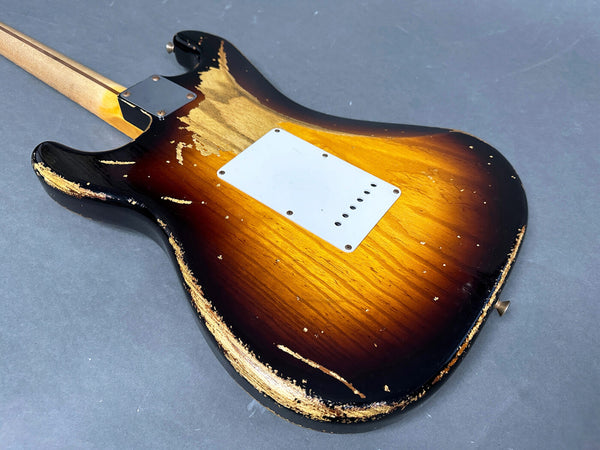 Sunburst electric guitar body with heavy relic wear, showing wood grain and a white backplate, laid on a gray surface.
