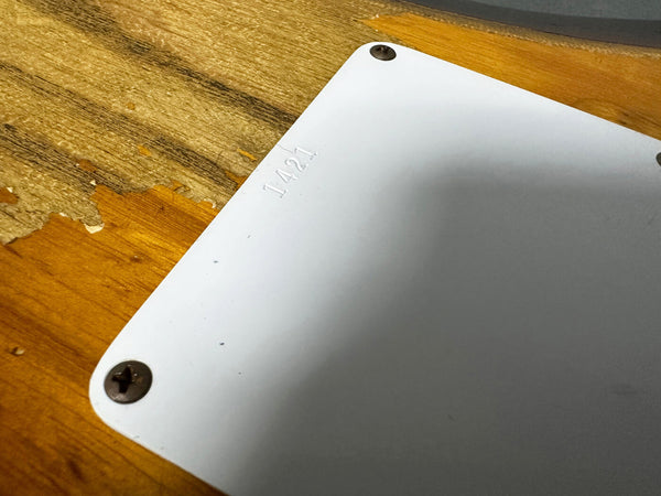 Close-up of a white plastic guitar backplate with embossed serial number 1421, mounted on a natural wood guitar body showing wear and scratches.
