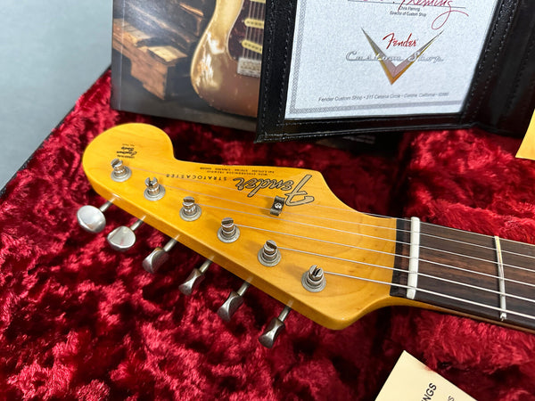 Close-up of Fender Stratocaster guitar headstock with tuning pegs, maple wood finish, and vintage-style logo, resting on red plush case lining with certificate from Fender Custom Shop visible in the background.