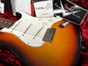 Close-up of sunburst Fender Stratocaster electric guitar body with white pickguard, three single-coil pickups, volume and tone knobs, chrome bridge, and included Fender Custom Shop and TUSQ packaging in red plush guitar case