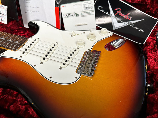 Close-up of sunburst Fender Stratocaster electric guitar body with white pickguard, three single-coil pickups, volume and tone knobs, chrome bridge, and included Fender Custom Shop and TUSQ packaging in red plush guitar case