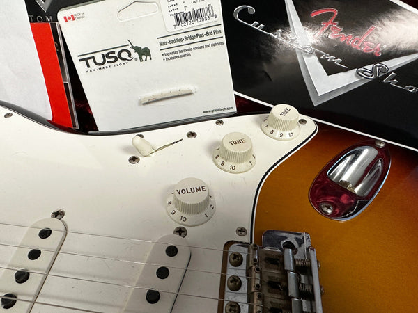 Close-up of vintage sunburst Fender Stratocaster electric guitar body with white pickguard, tone and volume knobs, and Tremolo bridge, alongside package of TUSQ man-made ivory guitar bridge pins and Fender custom shop paperwork.