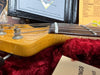 Close-up of a natural wood guitar headstock with tuning pegs and strings against a red plush lining, showing part of Fender Custom Shop documentation in the background