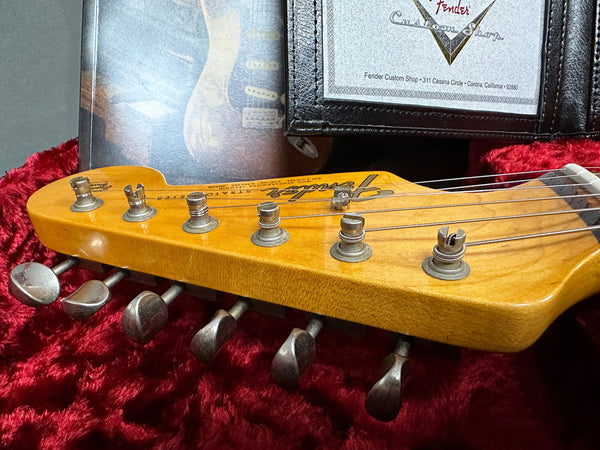 Close-up of vintage Fender Stratocaster guitar headstock with tuning pegs on red plush lining, accompanied by Fender Custom Shop certificate and booklet in the background.