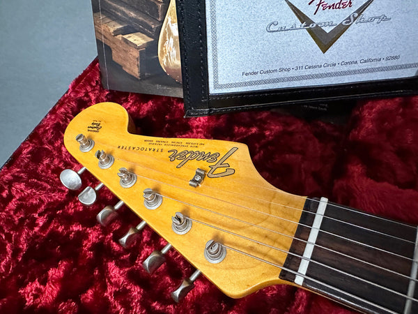 Close-up of Fender Stratocaster guitar headstock with tuning pegs and partial view of certificate on red velvet background