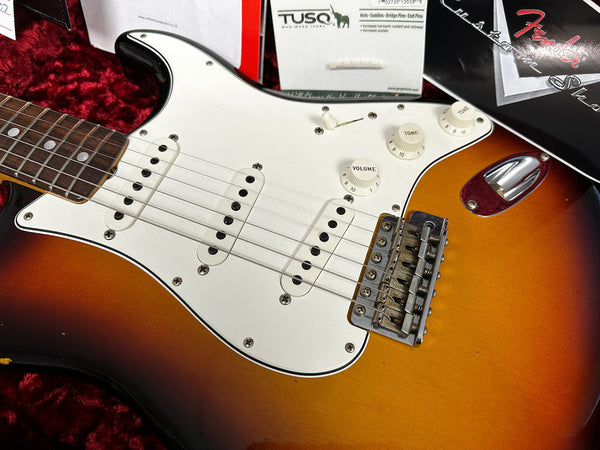 Close-up of sunburst Fender Stratocaster electric guitar body with white pickguard, three single-coil pickups, volume and tone control knobs, and Tremolo bridge, displayed in red velvet case with Fender and TUSQ branded packaging.