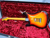 Vintage Fender Stratocaster electric guitar in sunburst finish shown from back, resting inside red velvet-lined guitar case with Fender Custom logo, showcasing worn finish and metal neck plate.