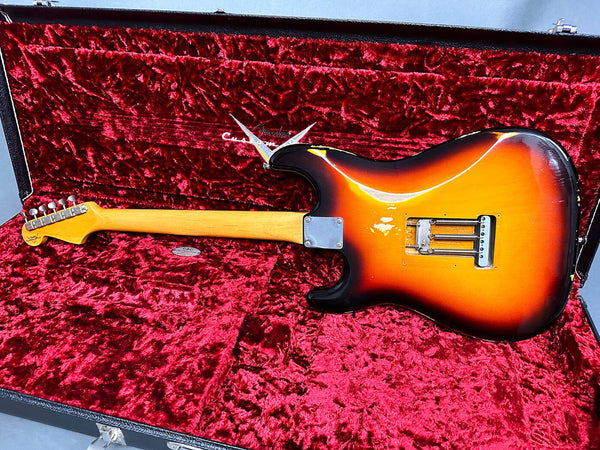Vintage Fender Stratocaster electric guitar in sunburst finish shown from back, resting inside red velvet-lined guitar case with Fender Custom logo, showcasing worn finish and metal neck plate.