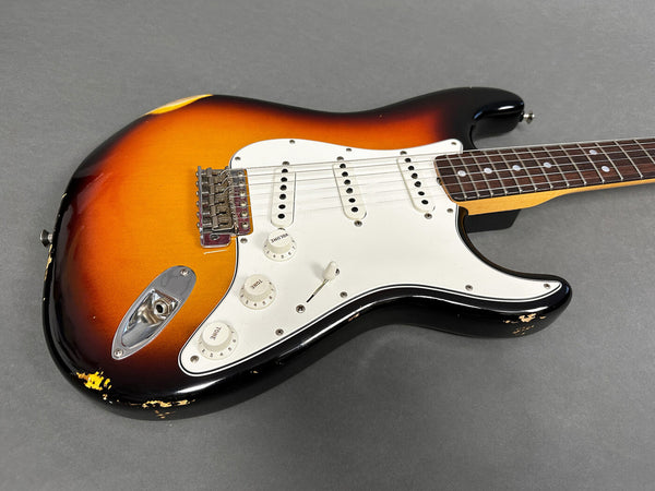Close-up of vintage sunburst electric guitar body with white pickguard, three single-coil pickups, and chrome bridge on gray background
