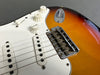 Close-up of sunburst electric guitar body showing white pickguard, three single-coil pickups, volume and tone control knobs, and chrome bridge saddle with six adjustable saddles