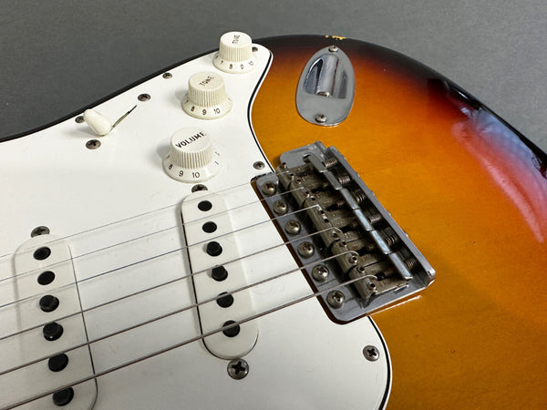 Close-up of sunburst electric guitar body showing white pickguard, three single-coil pickups, volume and tone control knobs, and chrome bridge saddle with six adjustable saddles