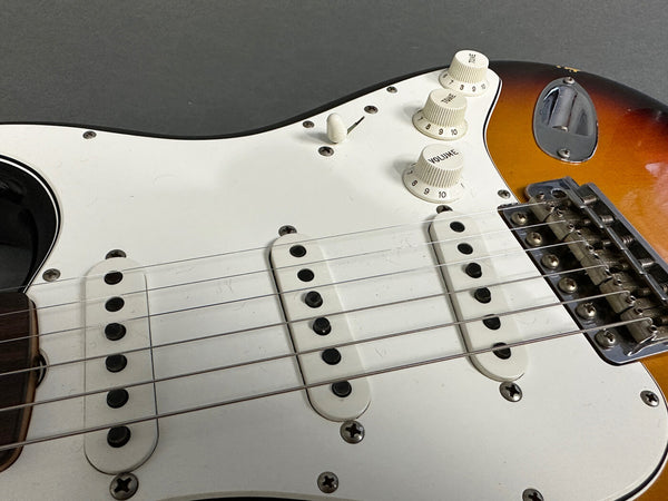 Close-up of electric guitar body with three single-coil pickups, white pickguard, tone and volume knobs, sunburst finish, and chrome bridge assembly