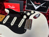 Close-up of white Fender electric guitar with black pickguard, three single-coil pickups, volume and tone knobs, vintage-style bridge, signed Fender Custom Shop certificate and manual in red and black case lining