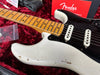 Close-up of vintage white Fender Stratocaster electric guitar with maple neck and black pickguard in red velvet case, showing wear and finish cracks