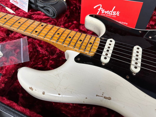 Close-up of vintage white Fender Stratocaster electric guitar with maple neck and black pickguard in red velvet case, showing wear and finish cracks