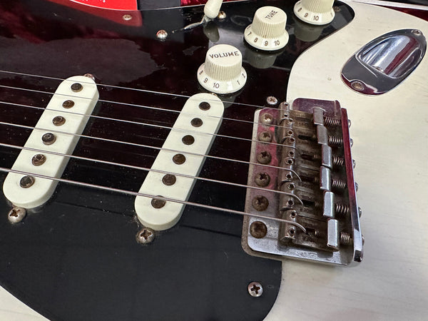 Close-up of electric guitar body showing white single-coil pickups, volume and tone control knobs, and metal bridge with guitar strings.