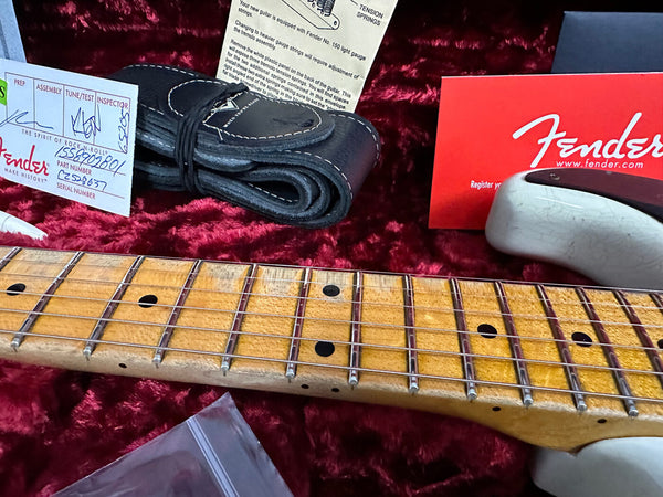 Close-up of Fender electric guitar maple neck with black dot inlays and metal frets, resting on red velvet lining in case, alongside black leather guitar strap, Fender certificate of authenticity, and Fender warranty card.