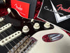 Close-up of vintage Fender Stratocaster electric guitar body featuring white volume and tone knobs, black pickguard, three single-coil pickups, chrome bridge, and Fender Custom Shop certificate in background.