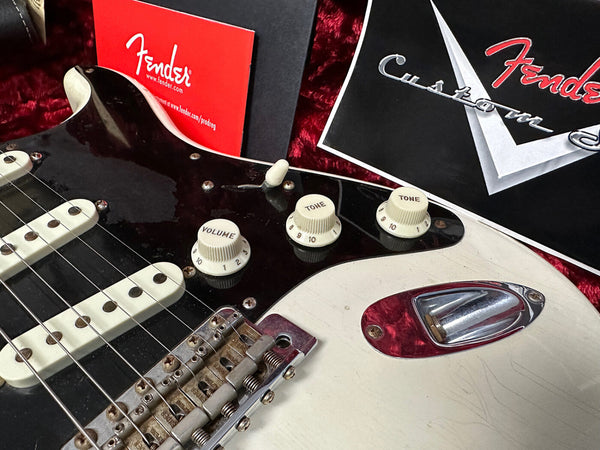 Close-up of vintage Fender Stratocaster electric guitar body featuring white volume and tone knobs, black pickguard, three single-coil pickups, chrome bridge, and Fender Custom Shop certificate in background.