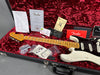 Fender electric guitar with worn white body and maple neck inside red plush-lined black hard case, accompanied by Fender certificate of authenticity, user manual, tremolo springs instruction sheet, leather strap, and various replacement parts in plastic bags.