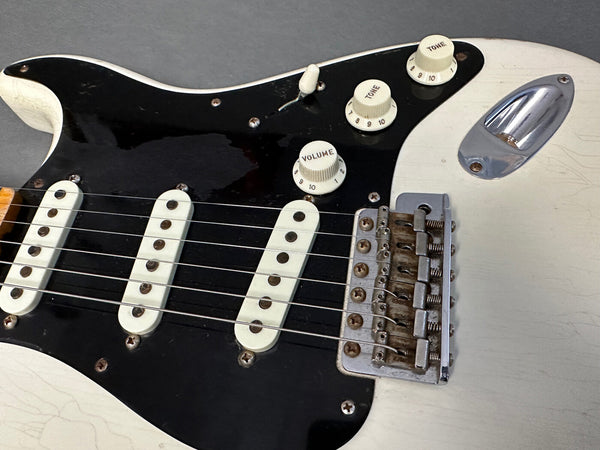 Close-up of vintage white electric guitar body showing black pickguard, three single-coil pickups, volume and tone knobs, and worn metal bridge assembly