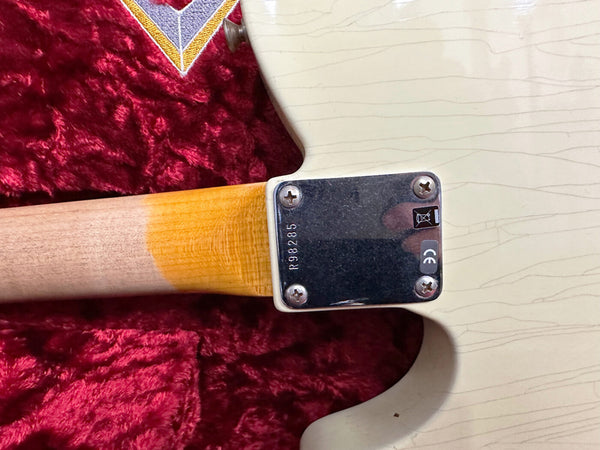 Close-up of electric guitar neck joint showing metal neck plate with screws, serial number R98285, maple neck wood, and cream body with visible finish checking on red plush case background