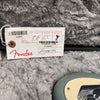 Fender guitar inspection tag with assembly, tune/test, inspector details, and part number VS17081191 on gray plush background next to electric guitar body edge