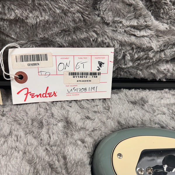 Fender guitar inspection tag with assembly, tune/test, inspector details, and part number VS17081191 on gray plush background next to electric guitar body edge