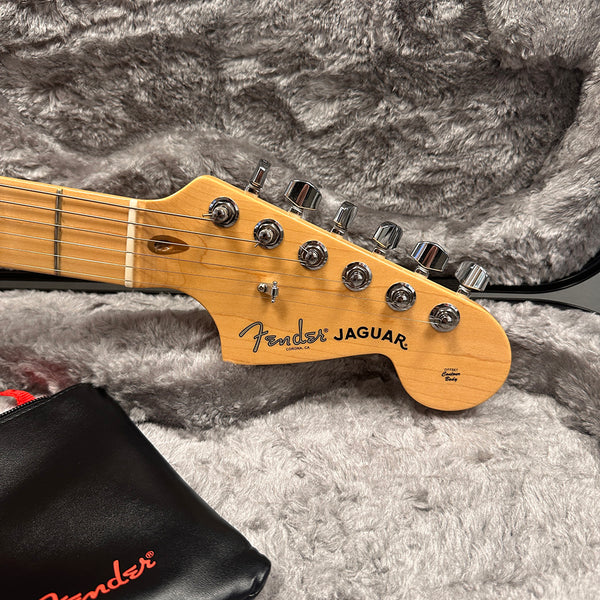 Fender Jaguar electric guitar headstock with chrome tuning pegs and Fender logo, resting on plush gray case lining with black Fender accessory pouch.