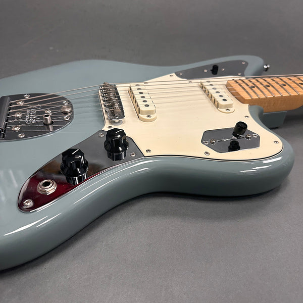 Close-up of light blue electric guitar body with white pickguard, two single-coil pickups, black control knobs, and light maple neck on gray background