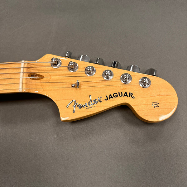 Close-up of natural wood Fender Jaguar guitar headstock with chrome tuning pegs and black logo text on gray background