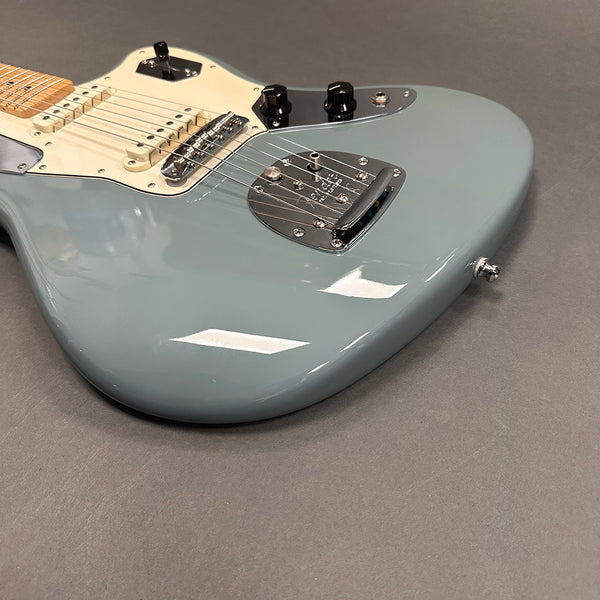 Close-up of a light blue electric guitar body with cream pickguard, black control knobs, and chrome bridge and tailpiece on a gray surface.