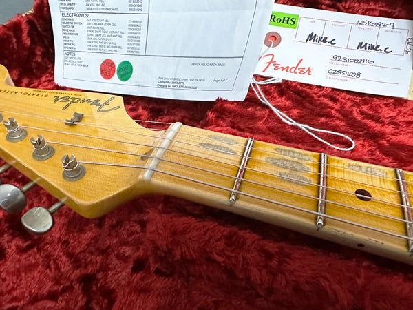 Close-up of Fender Stratocaster guitar headstock and fretboard with tuning pegs on red plush guitar case interior showing Fender authenticity tag and electronics specification sheet.