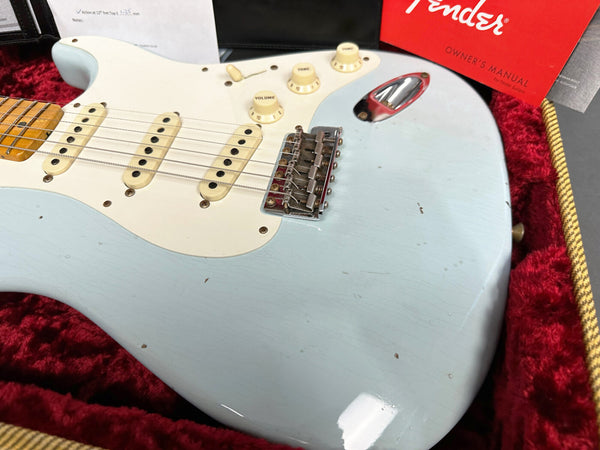Close-up of vintage light blue Fender Stratocaster electric guitar body showing cream pickguard, three single-coil pickups, volume and tone knobs, and chrome bridge, resting in red velvet-lined guitar case with Fender owner's manual in background