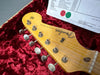 Close-up of Fender Stratocaster guitar headstock with vintage tuning pegs, natural wood finish, and certification paper in red velvet-lined case