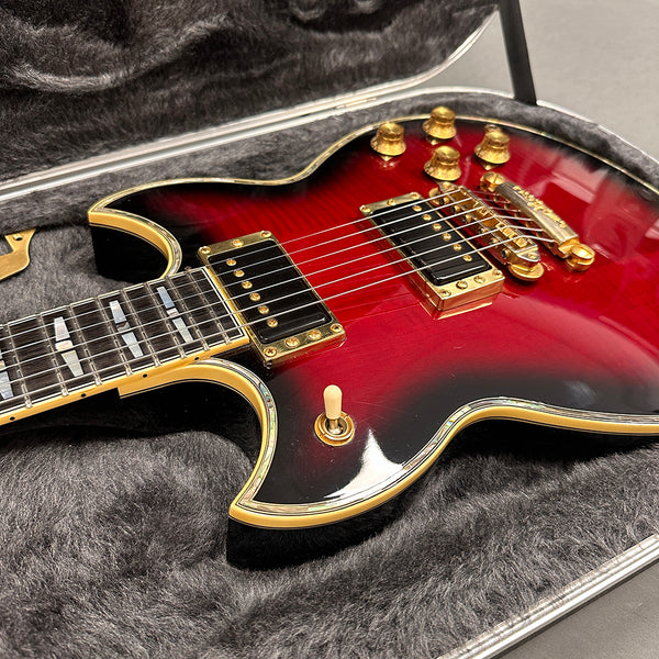 Close-up of red and black electric guitar with gold hardware and dual humbucker pickups resting in a gray plush-lined hard case