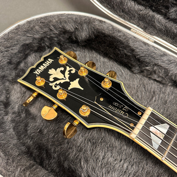 Close-up of Yamaha Custom SG 3000 guitar headstock with gold tuning pegs and floral inlay inside a plush gray guitar case