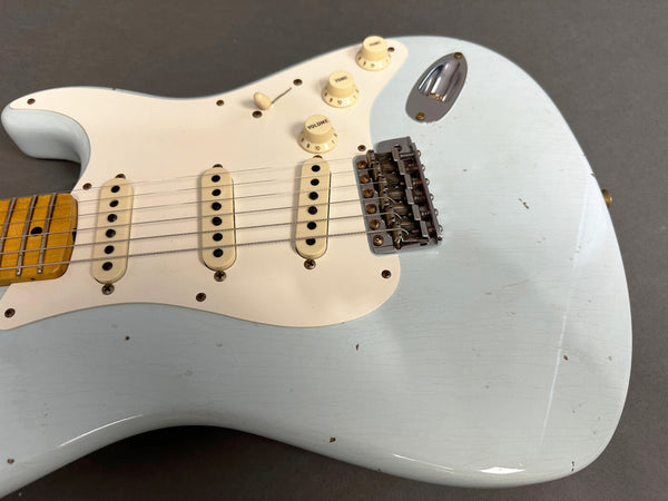 Close-up of light blue electric guitar body with white pickguard, three single-coil pickups, volume and tone knobs, and vintage-style bridge showing wear and aging details