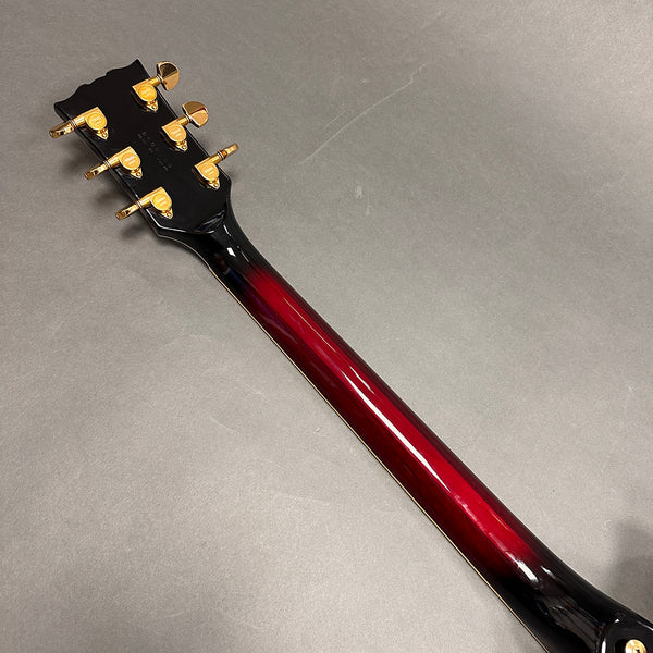 Back view of guitar neck with red to black gradient finish and gold tuning pegs on gray background