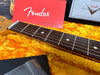 Close-up of Fender guitar neck with rosewood fretboard, dot inlays, and steel strings, alongside Fender owner's manual and Fender Custom Shop certificate in yellow plush guitar case lining.
