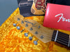 Close-up of vintage Fender Telecaster headstock with tuning pegs and strings, resting on bright yellow guitar case lining, with worn Fender Stratocaster body and red Fender owner's manual in the background.