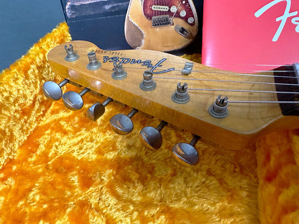 Close-up of vintage Fender Telecaster guitar headstock with tuning pegs, resting on orange plush guitar case interior, with partial view of Fender Stratocaster guitar and red Fender box in the background.