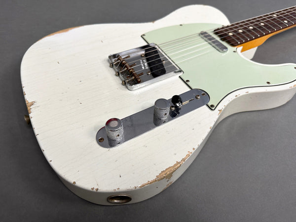 Close-up of vintage white electric guitar body with worn finish, chrome bridge, control knobs, and mint green pickguard on gray background