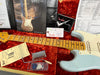 Fender electric guitar with maple neck and light blue body resting in red velvet-lined case, surrounded by Fender Custom Guitar design guide, tremolo springs instruction card, tremolo arm, guitar picks, Fender documentation, and certificate of authenticity.