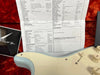 Close-up of Coffee House Guitars instrument details sheet for 56 Strat guitar model with specifications including body style, pickup route, wood types, hardware components, and electronics, partially visible light blue vintage-style guitar body on red plush background.