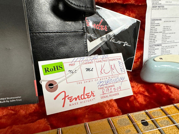 Fender guitar tag with serial number and part number, RoHS compliance label, and certificate card inside red plush guitar case with maple fretboard and blue guitar body edge visible