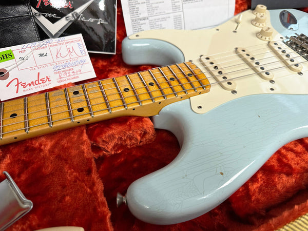 Close-up of a vintage Fender electric guitar with cracked light blue body and aged maple neck, resting in an orange plush guitar case alongside Fender documentation and inspection tags.