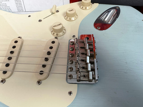 Close-up of light blue electric guitar body showing white pickguard, volume and tone control knobs, two single-coil pickups, and chrome bridge with Fender engraved saddles