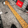 Close-up of a Fender electric bass guitar neck with maple fingerboard and chrome tuning pegs resting in a gray plush-lined hard case with Fender owner's manual and accessories.