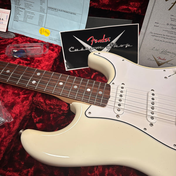Close-up of white Fender Stratocaster electric guitar body and rosewood fretboard on red plush guitar case with Fender Custom Shop certificate and paperwork background